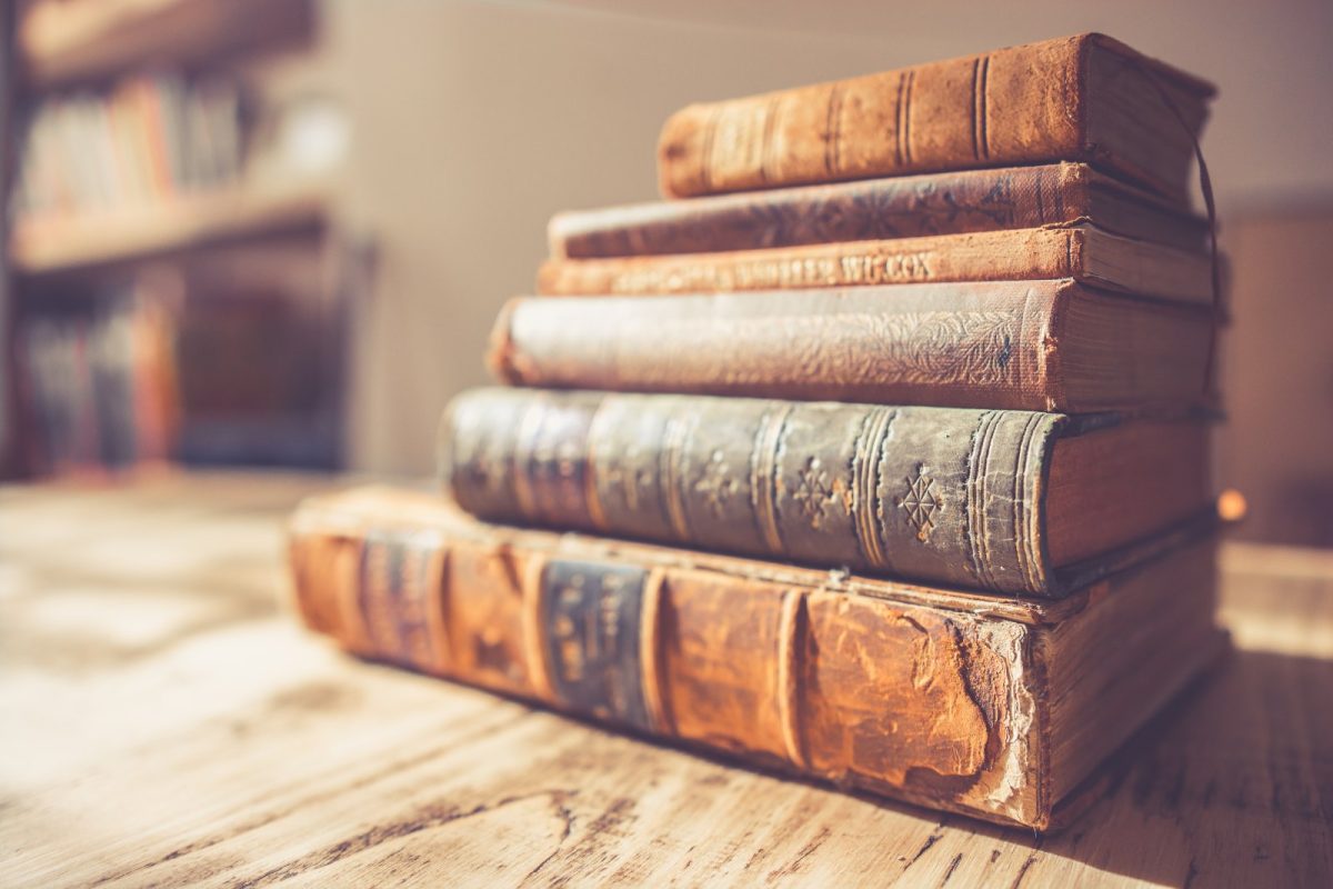 Pila de libros antiguos de tapa de cuero sobre una mesa de madera, con una estantería desenfocada al fondo