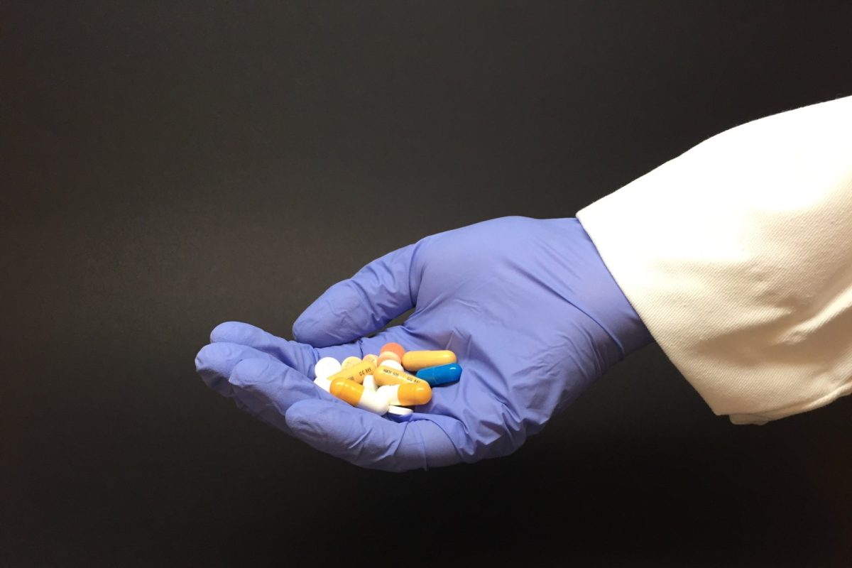 Mano con guante médico azul y bata blanca sosteniendo un puñado de pastillas y cápsulas de colores variados sobre fondo oscuro