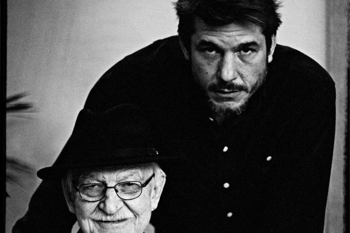 Antonio Escohotado sonriente, sentado, con sombrero y gafas, en un retrato en blanco y negro junto a su hijo Jorge
