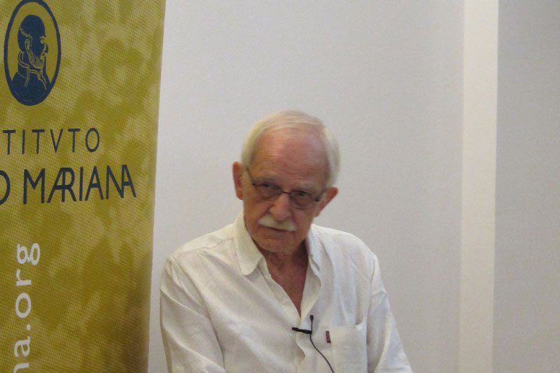 Antonio Escohotado, de pelo blanco, gafas y bigote, sentado y hablando en una conferencia