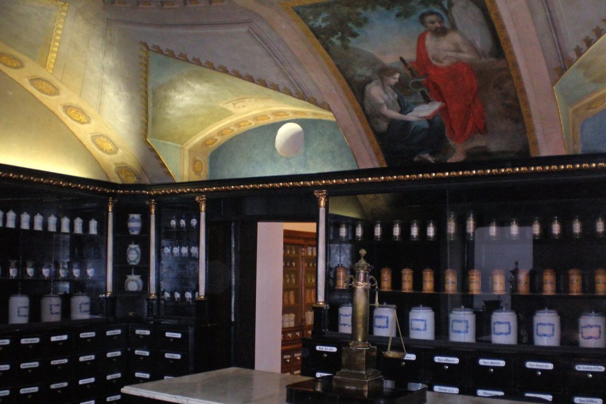 Sala de un antiguo museo de farmacia en Bratislava con estanterías de botes de farmacia de cerámica blanca y azul, columnas doradas, una balanza de latón sobre un mostrador de mármol y un techo abovedado pintado con frescos