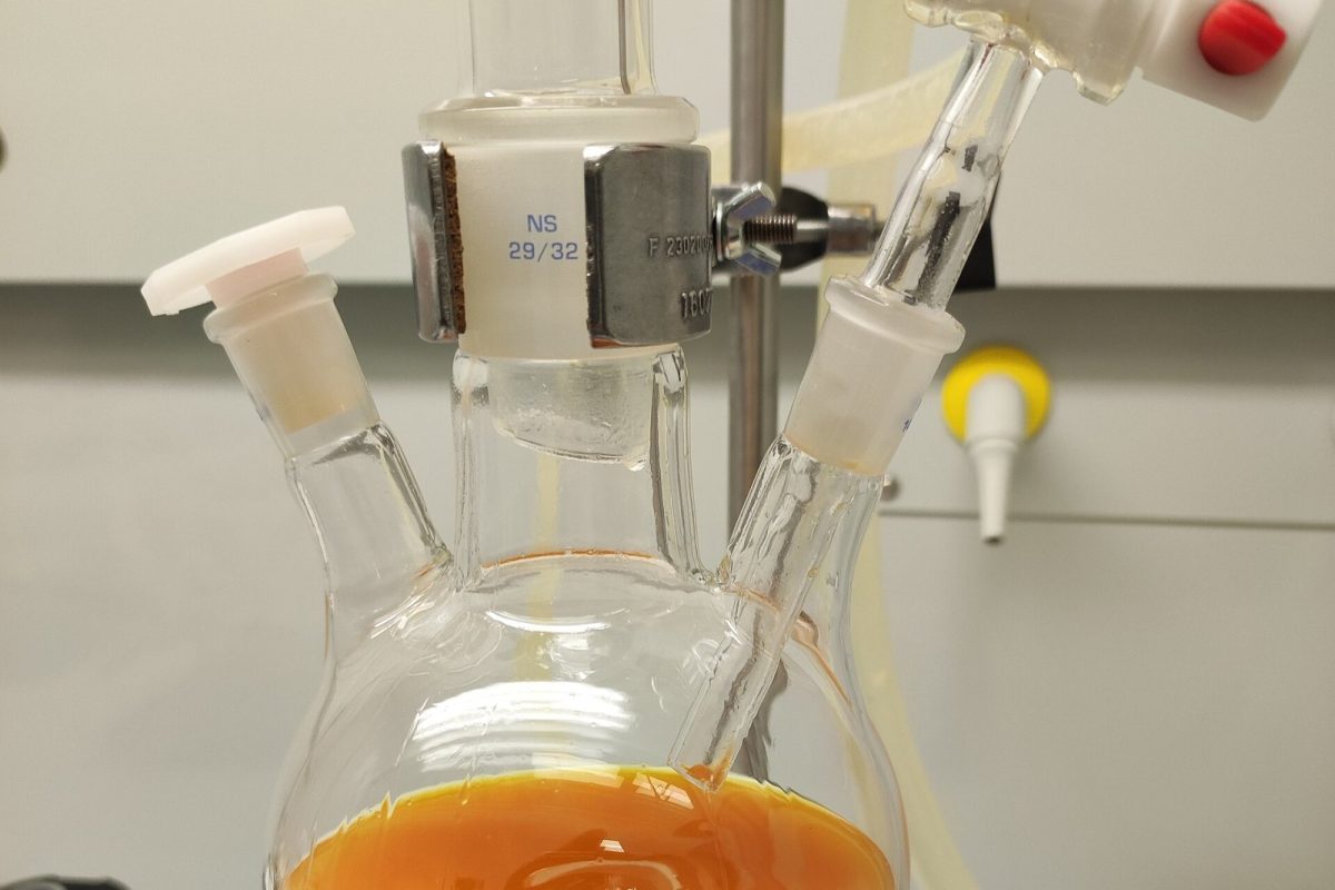 Matraz tricuello de laboratorio con un líquido de color naranja intenso en plena reacción, montado sobre una placa agitadora