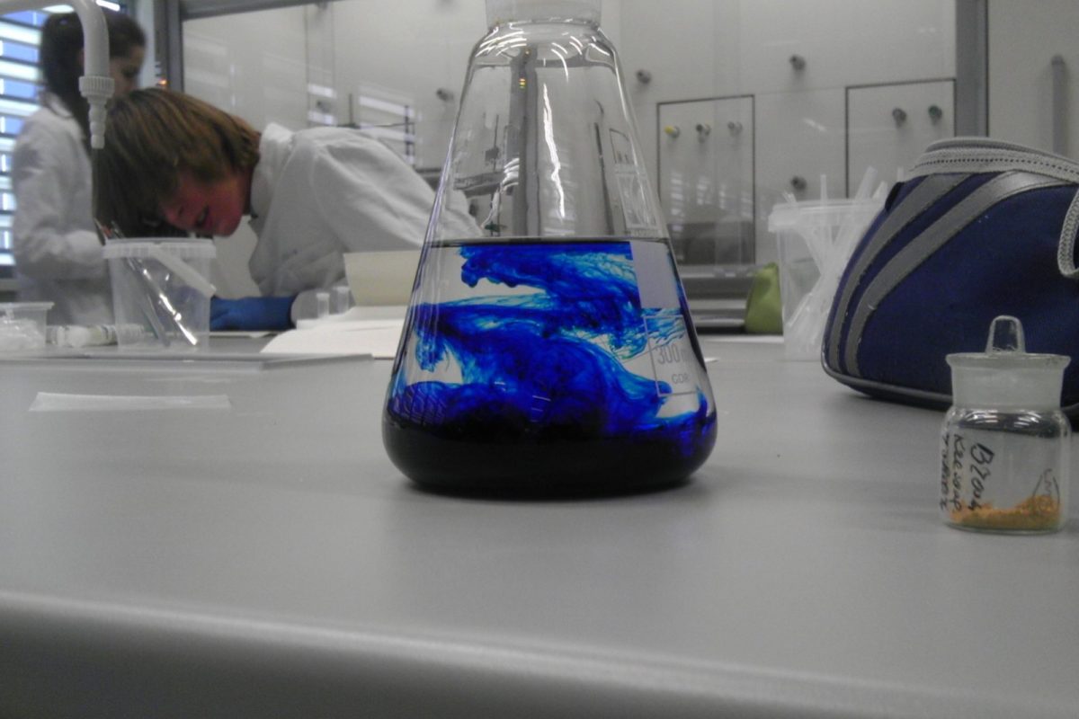 Matraz Erlenmeyer con un colorante azul difuminándose en el líquido, con personal de laboratorio al fondo