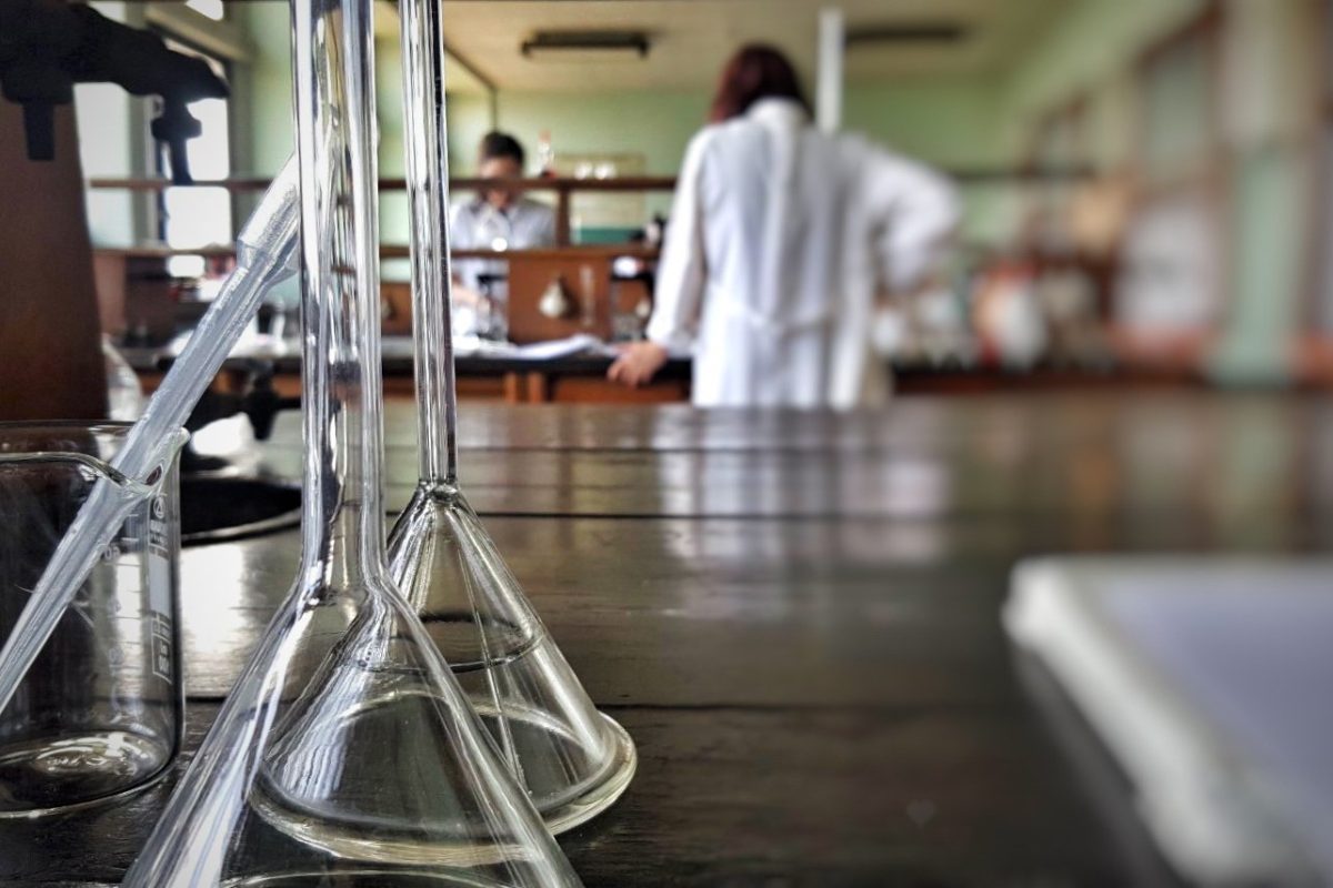Matraces y vasos de precipitado en primer plano sobre una mesa de laboratorio, con estudiantes de bata blanca al fondo