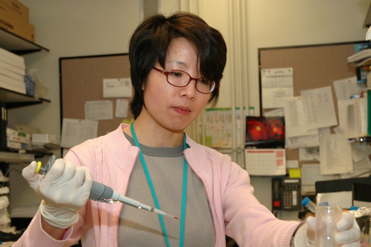 Científica con guantes y bata de laboratorio utilizando una micropipeta para transferir una muestra a un matraz, en un laboratorio de análisis.