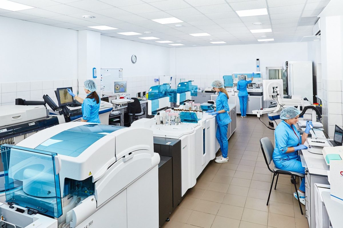 Varios técnicos con bata y gorro sanitario trabajando con analizadores automáticos en un laboratorio clínico moderno y luminoso.