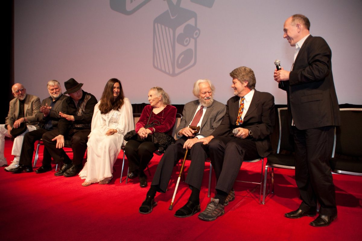Alexander Shulgin, de edad avanzada y apoyado en un bastón blanco, sentado junto a otros invitados en un panel sobre un escenario con alfombra roja