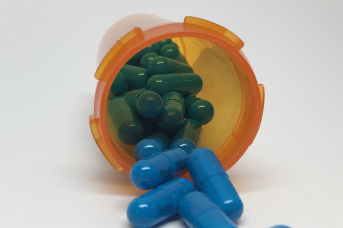Cápsulas azules y verdes cayendo de un bote de medicamento naranja sobre una superficie blanca