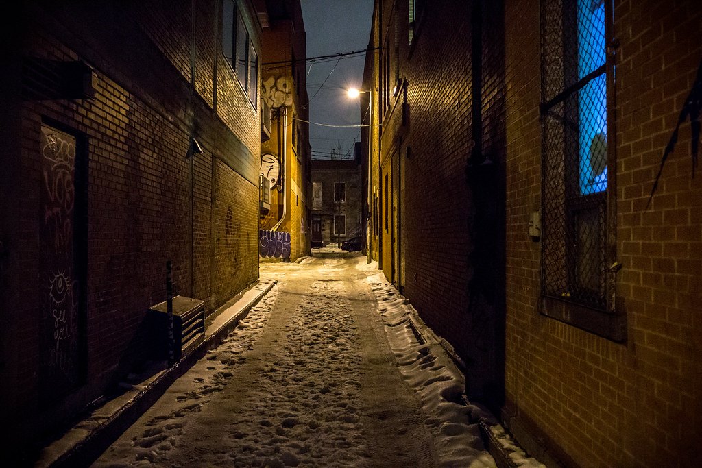 Callejón estrecho y oscuro entre edificios de ladrillo, con nieve en el suelo y una farola encendida al fondo