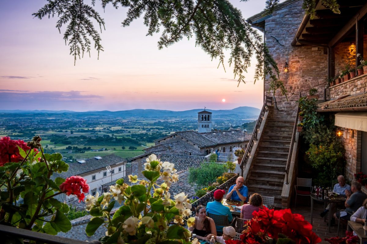 Personas cenando en una terraza con vistas a un valle italiano al atardecer, con flores en primer plano
