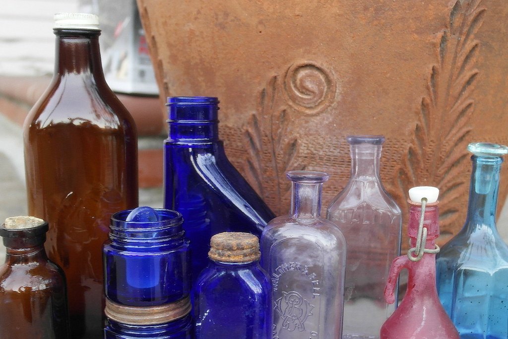 Colección de antiguos frascos de vidrio de farmacia en tonos ámbar, azul y transparente, agrupados sobre una superficie de piedra.