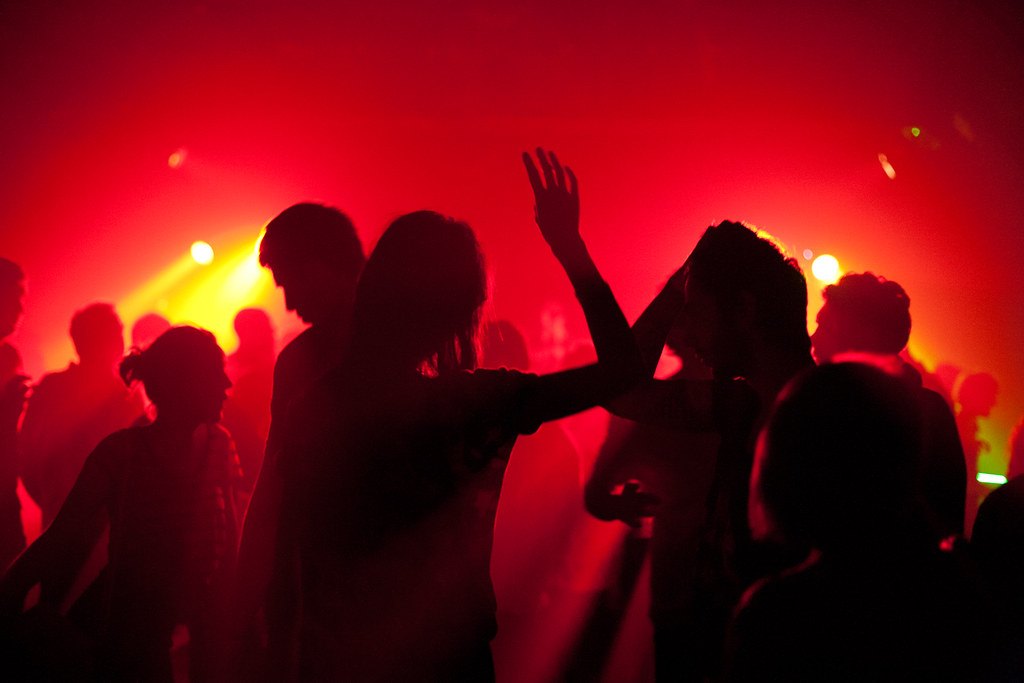 Siluetas de personas bailando y levantando los brazos en una discoteca envuelta en luces rojas y doradas.