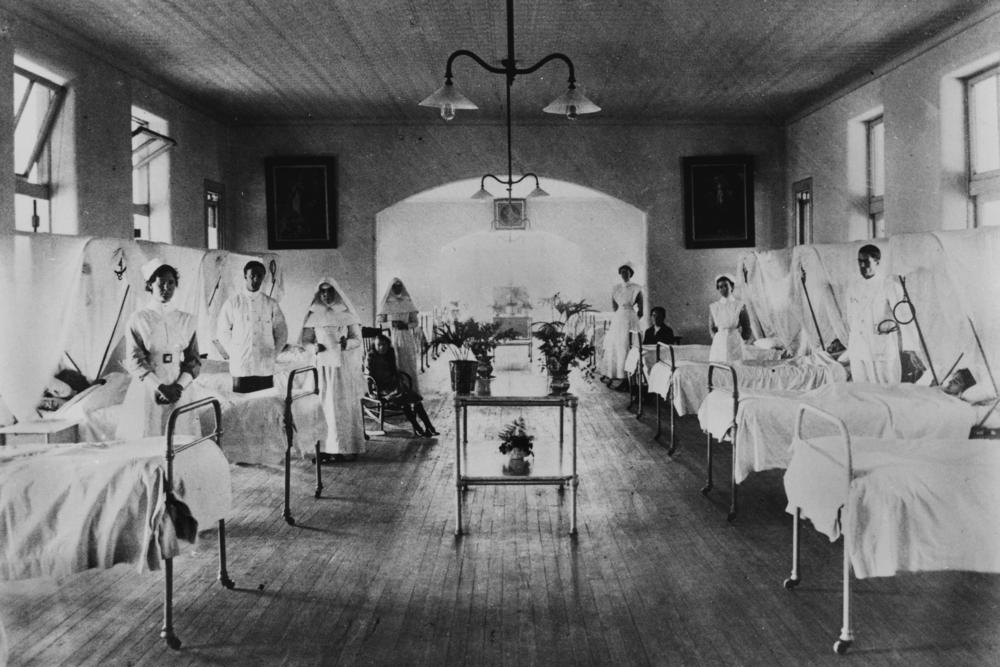 Fotografía en blanco y negro de una sala de hospital antigua con camas en fila y personal de enfermería de uniforme, año 1914