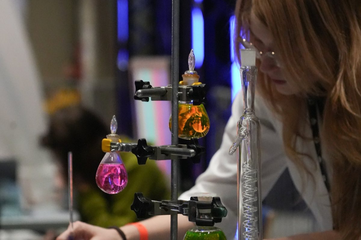 Matraces de laboratorio con líquidos de colores vivos montados en un soporte metálico, con una persona de bata blanca trabajando al fondo