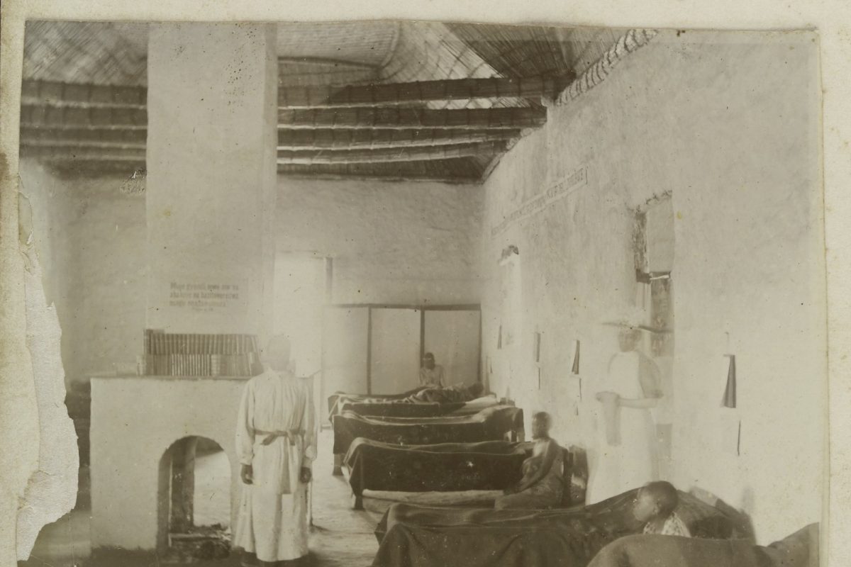 Fotografía antigua en blanco y negro de una sala de hospital con pacientes en cama y una enfermera de pie, con la inscripción manuscrita "A bit of the Women's Ward"