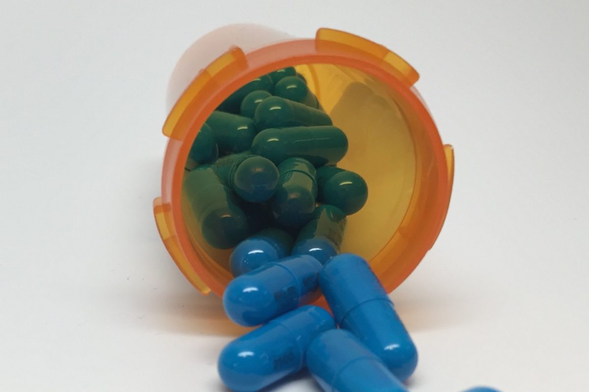 Cápsulas verdes y azules cayendo de un bote de medicamento con tapa naranja.