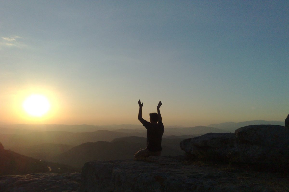 Silueta de una persona en actitud meditativa con los brazos alzados frente a un atardecer en la montaña