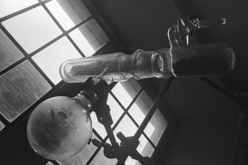 Fotografía en blanco y negro de un matraz y un aparato de destilación de vidrio junto a una ventana en un laboratorio