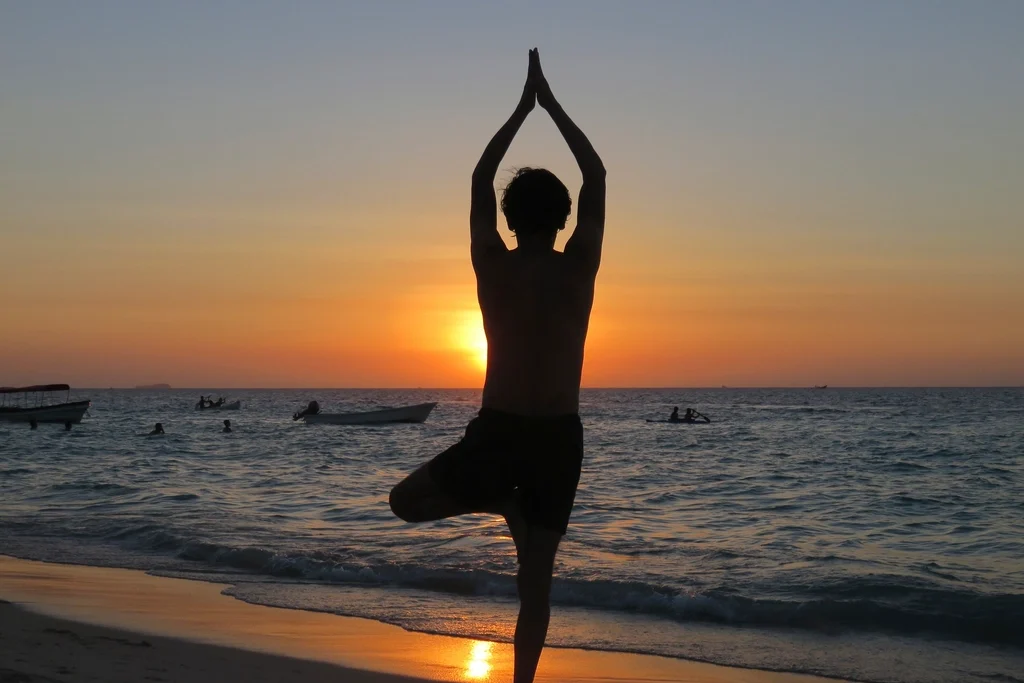 Silueta de una persona en postura de árbol de yoga sobre una playa, con el sol poniéndose sobre el mar al fondo
