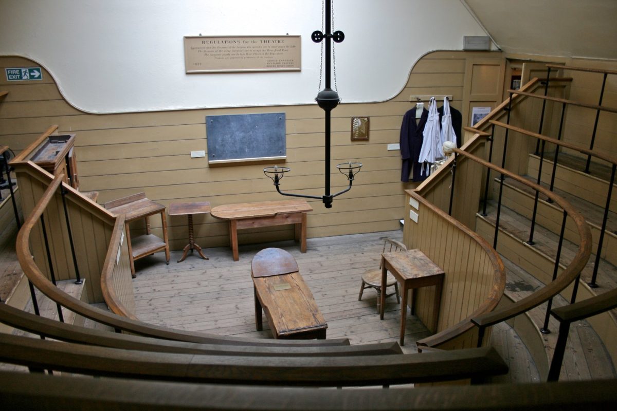 Interior del histórico Old Operating Theatre de Londres: un antiguo quirófano de madera con mesa de operaciones central, candelabro y graderío curvo para estudiantes, bajo el cartel de normas de 1822.