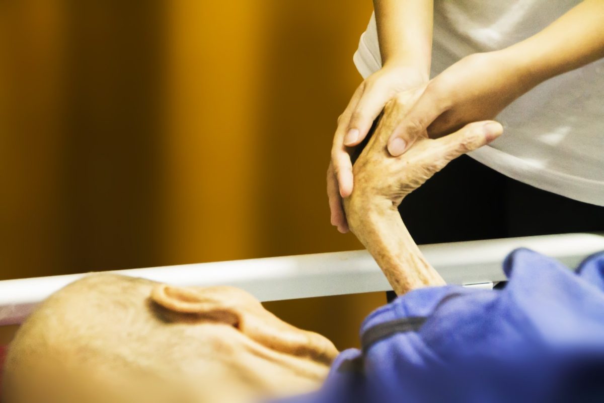 Las manos de una cuidadora sostienen con delicadeza la mano de un paciente anciano y frágil encamado en una unidad de cuidados paliativos.
