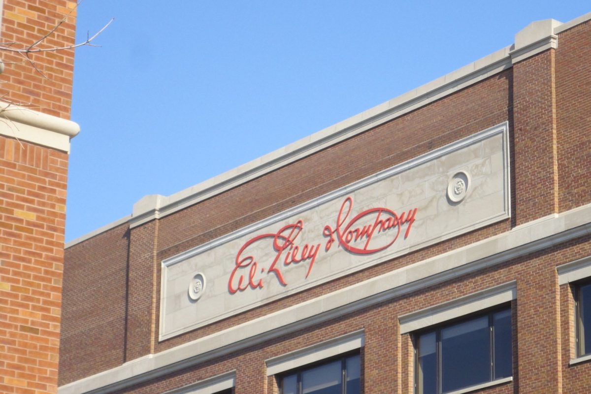 Fachada de ladrillo de un edificio de Eli Lilly and Company con el logotipo caligráfico de la empresa en rojo sobre una placa de piedra clara, bajo un cielo azul.