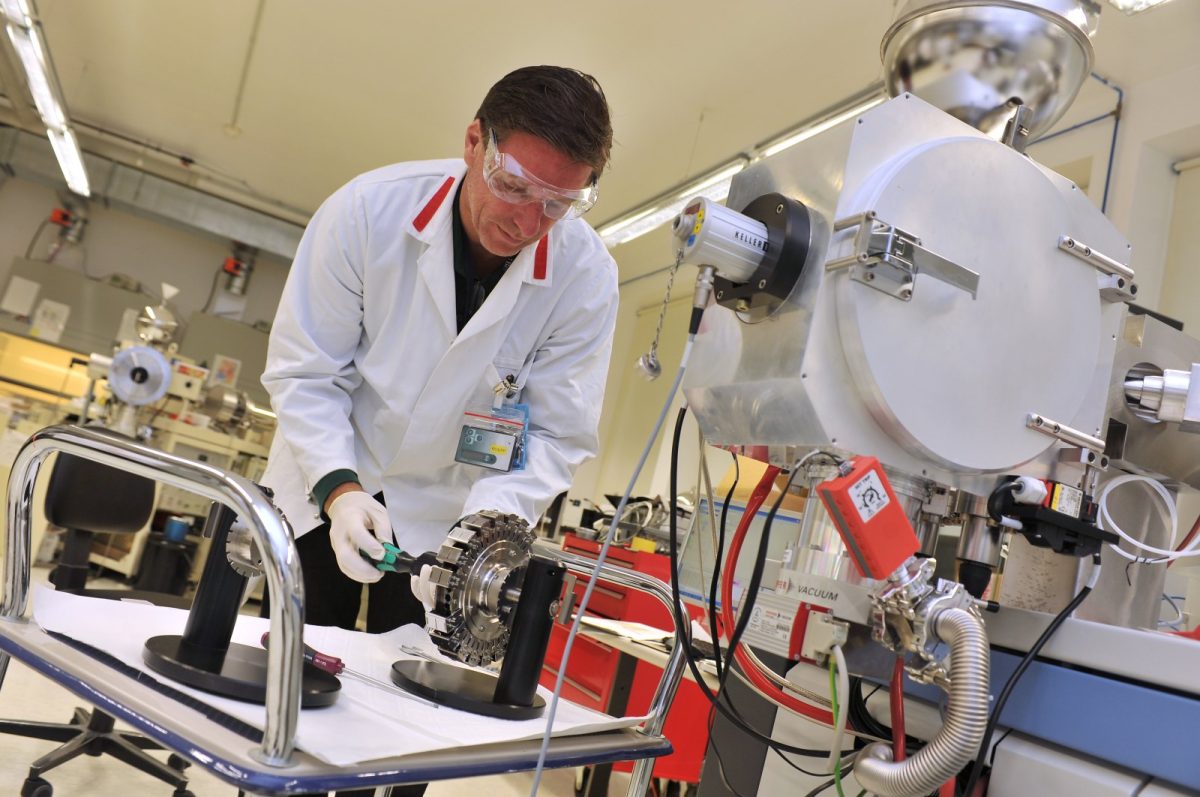 Un técnico con bata blanca y gafas de protección manipula el equipo de un laboratorio de espectrometría de masas.