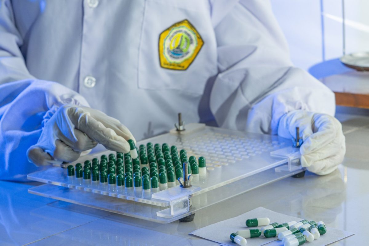 Trabajador de un laboratorio farmacéutico, con bata y guantes, colocando cápsulas verdes y blancas en una placa de llenado