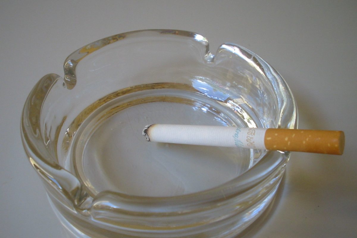 Un cigarrillo recién encendido apoyado sobre el borde de un cenicero de cristal transparente, sobre una superficie clara.
