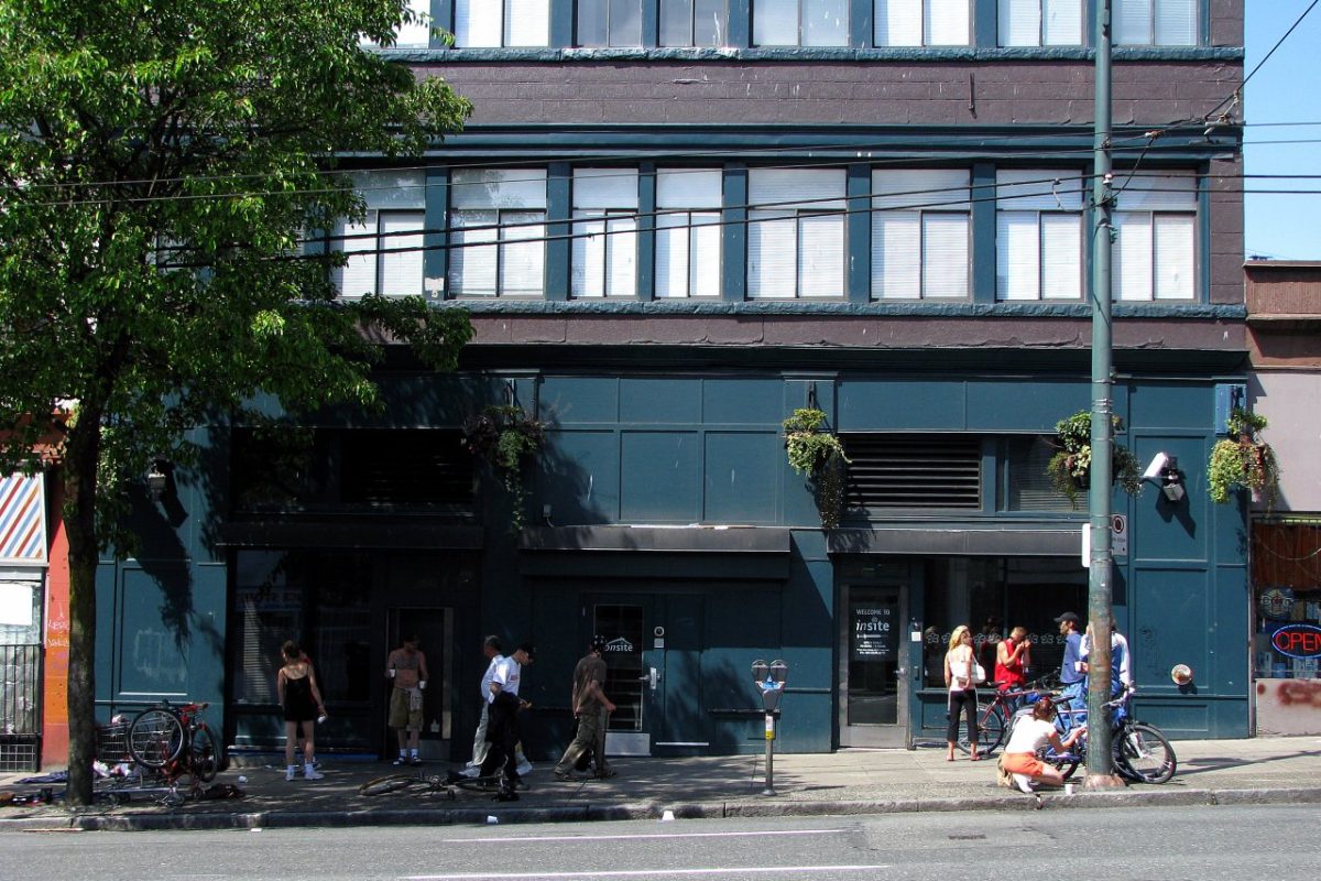 Fachada verde oscuro de un edificio con la entrada de Insite, una sala de consumo supervisado, y varias personas y bicicletas en la acera